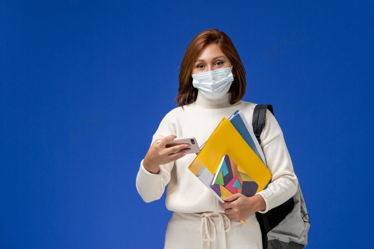 人正面图身穿白色球衣的年轻女学生戴着带包的面具和复印件 在蓝色墙上用手机风景使用课程
