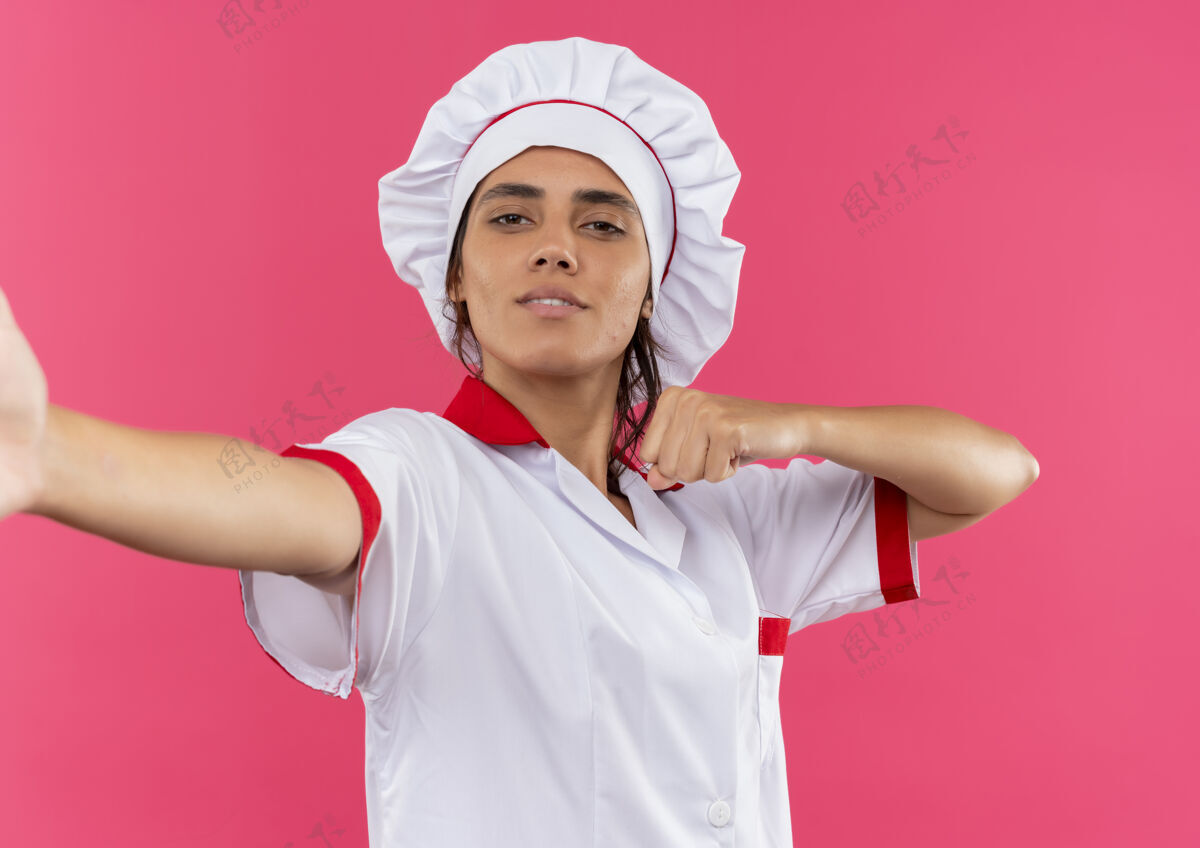 看年轻的女厨师穿着厨师制服 拿着相机在孤立的粉红色墙上展示拳头制服女拳头