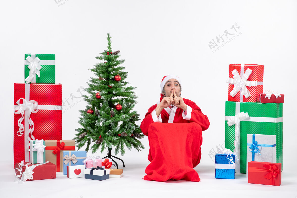 沉默情绪激动的年轻人装扮成圣诞老人 拿着礼物和装饰好的圣诞树 在白色背景上做着沉默的手势手势打扮兴奋