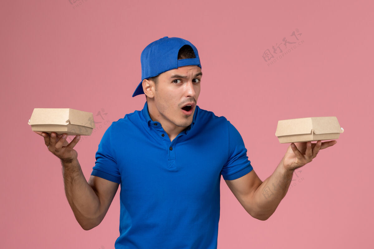 视图正面图：身穿蓝色制服披肩的年轻男性快递员在粉红色的墙上拿着小包裹持有食品年轻男性快递