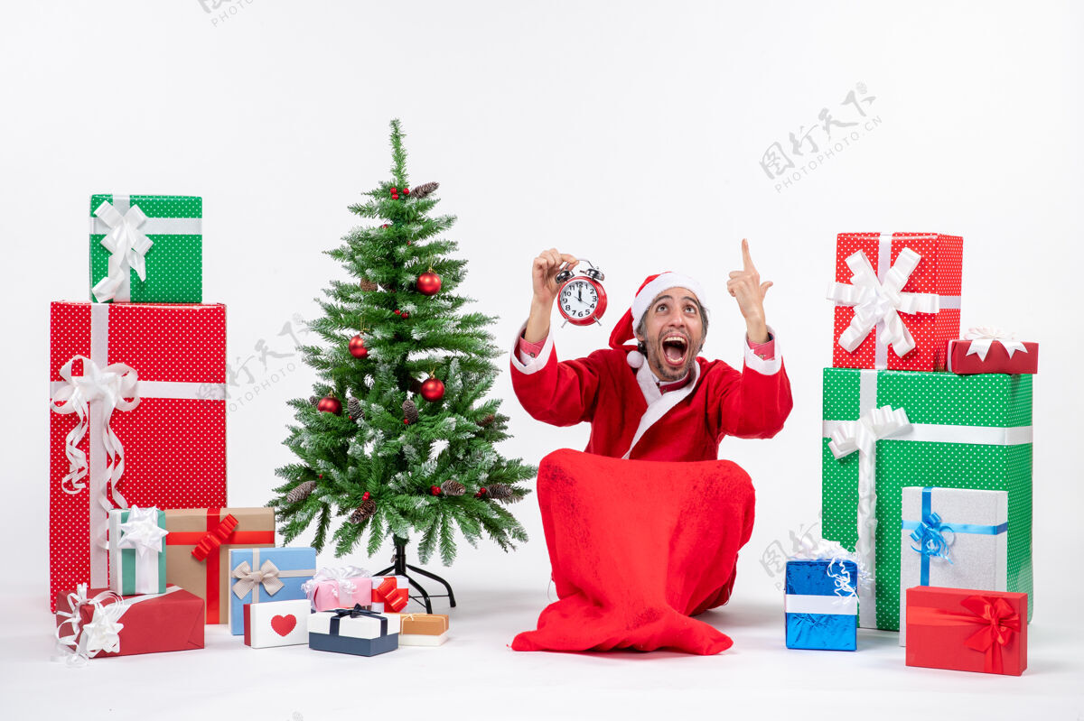 礼物紧张的圣诞老人坐在地上 在礼物和装饰圣诞树旁的白色背景上举起闹钟季节礼物盒子
