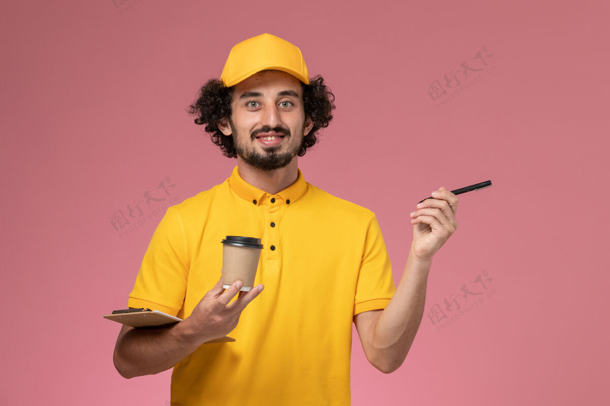 成人正面图：身穿黄色制服和披风的男性快递员拿着咖啡杯笔和笔记本 放在浅粉色的墙上专业送货帅哥