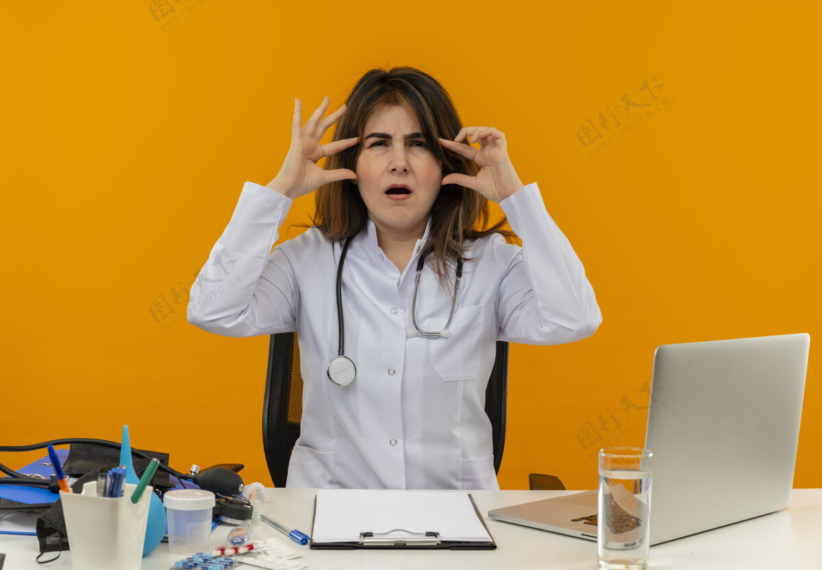 工具困惑的中年女医生穿着医用长袍 手持听诊器 坐在办公桌旁 手持医疗工具 将手指放在额头上 放在隔离的橙色墙壁上 留有复印空间工作手指长袍