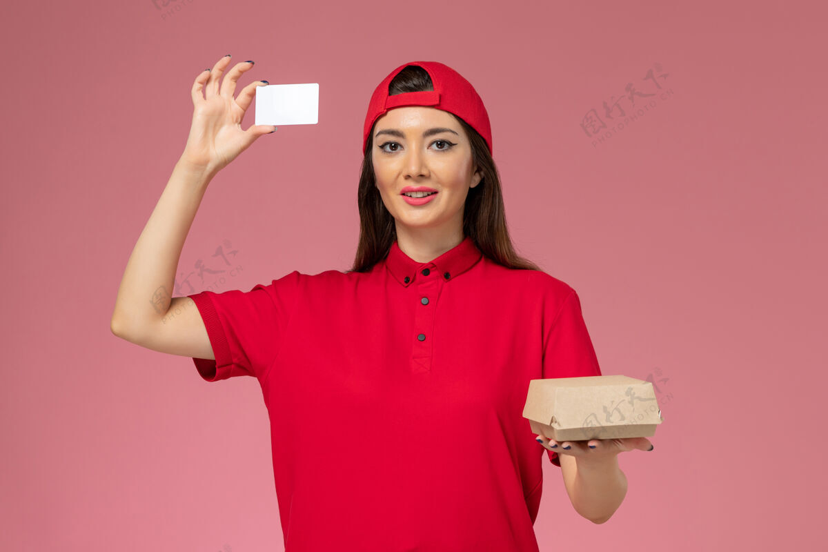 快乐正面图：身穿红色制服斗篷的年轻女快递员 手上拿着小小的快递食品包和卡片 放在浅粉色的墙上生意肖像食物