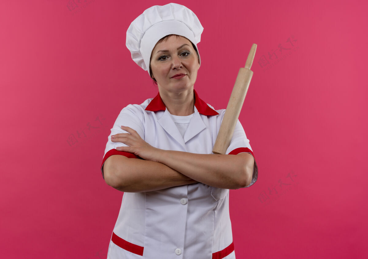拿着自信的中年女厨师穿着厨师制服 手拿擀面杖交叉在孤立的粉红色墙上手滚动厨师