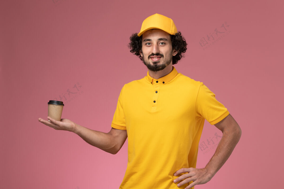 服务正面图：身穿黄色制服 披风的男性快递员在粉色墙上拿着送货咖啡杯男信使送货人