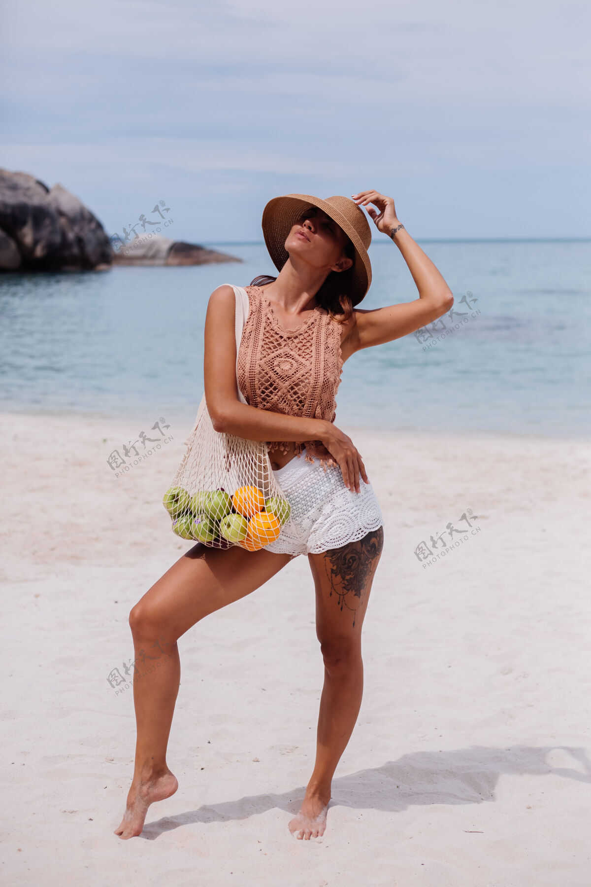 快乐年轻漂亮的白种人 皮肤黝黑 穿着针织服装 戴着草帽 手拿一个装满水果 苹果 橘子 葡萄的线袋 在热带海滩上手提包成人款式