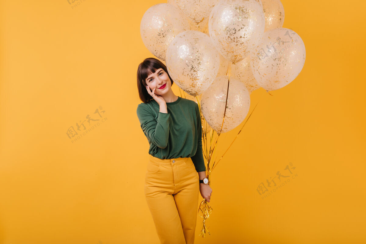 休闲迷人的直发女人摆出一束闪闪发光的气球室内拍摄的微笑无忧无虑的女孩在绿色毛衣和黄色裤子放松人魅力
