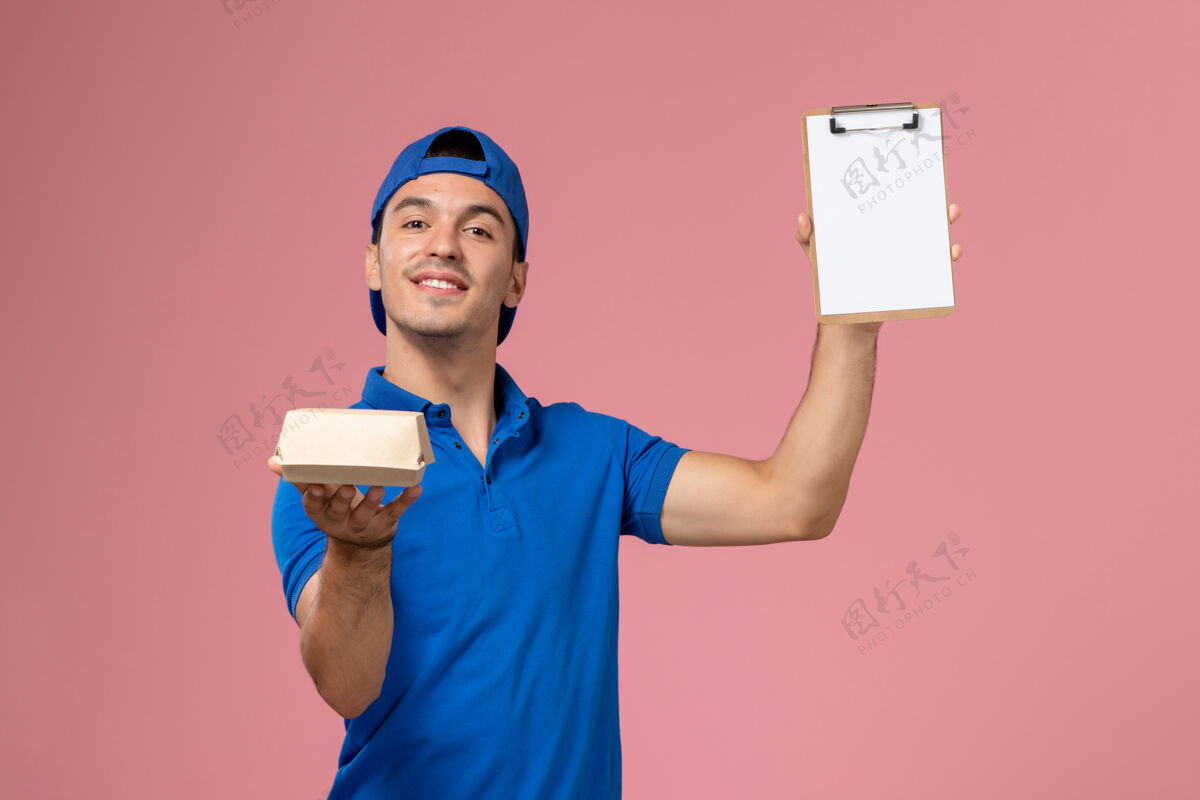 服务正面图身穿蓝色制服披肩的年轻男性快递员手持小快递食品包和笔记本在浅粉色的墙上正面人工人