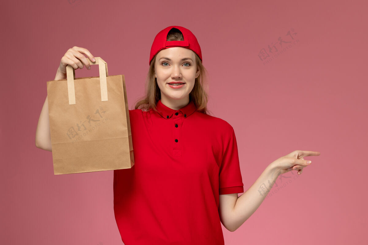 服务正面图：身穿红色制服 披风的年轻女快递员手持送食品的包裹 粉色墙上挂着微笑持有人肖像