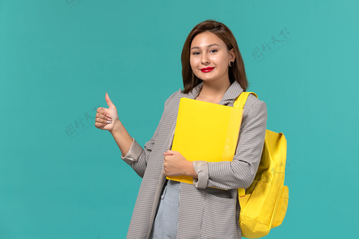 书籍身穿灰色夹克 背着黄色背包 在浅蓝色墙上拿着文件的女学生的正视图夹克女学生文件