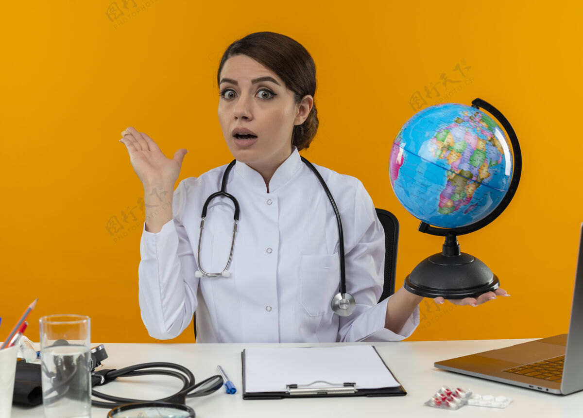 电脑关心的年轻女医生穿着医用长袍 手持听诊器 坐在办公桌旁 拿着医疗工具 拿着地球仪 用手指着隔离的黄色墙壁女医疗点