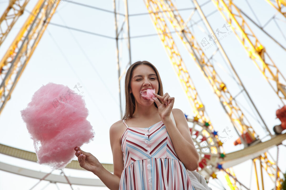情绪在温暖的日子里 一位穿着夏装的年轻美女站在游乐场的摩天轮旁 手里拿着棉花糖 嘴里塞着一块棉花糖 眨着眼睛户外20多岁夏天
