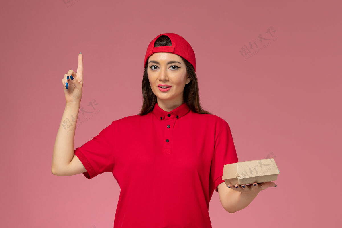粉色正面图：身穿红色制服和斗篷的年轻女快递员 手上拿着一个小小的快递食品包 放在粉红色的墙上正面工作制服