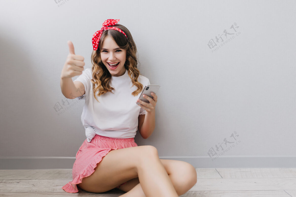 休闲可爱的白人女孩 戴着红丝带 黑发 坐在家里的地板上室内拍摄的是穿着粉色短裤微笑的女模特人电话室内