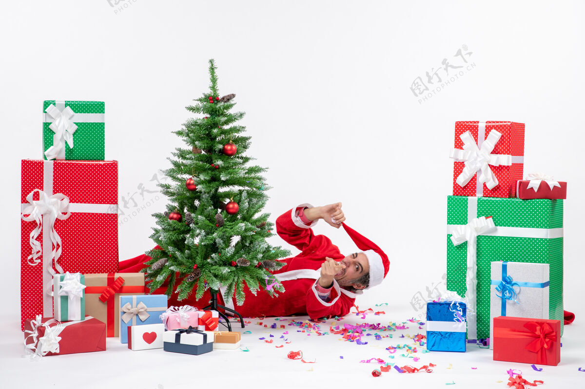 圣诞喜庆的节日气氛与年轻有趣的圣诞老人躺在圣诞树后附近的礼物白色背景股票照片圣诞老人车站圣诞老人