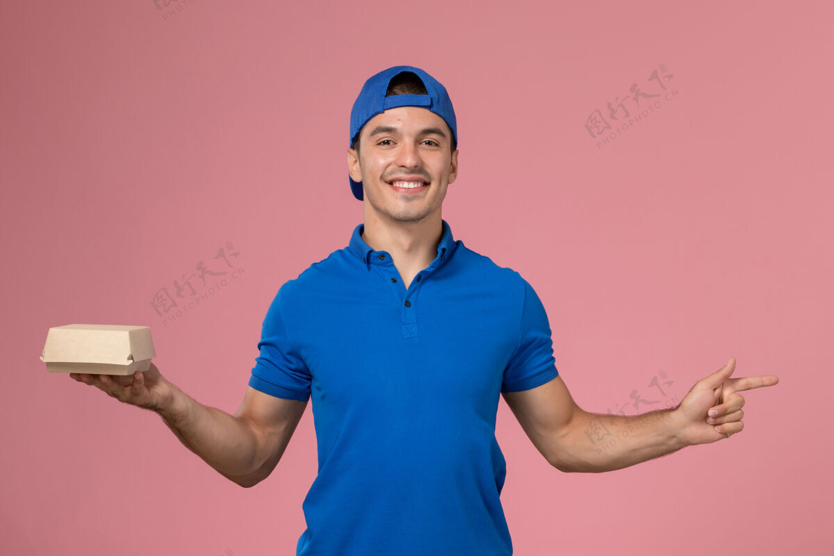 快递正面图：身穿蓝色制服披肩的年轻男性快递员手拿着小快递食品包站在粉红色的墙上制服正面肖像