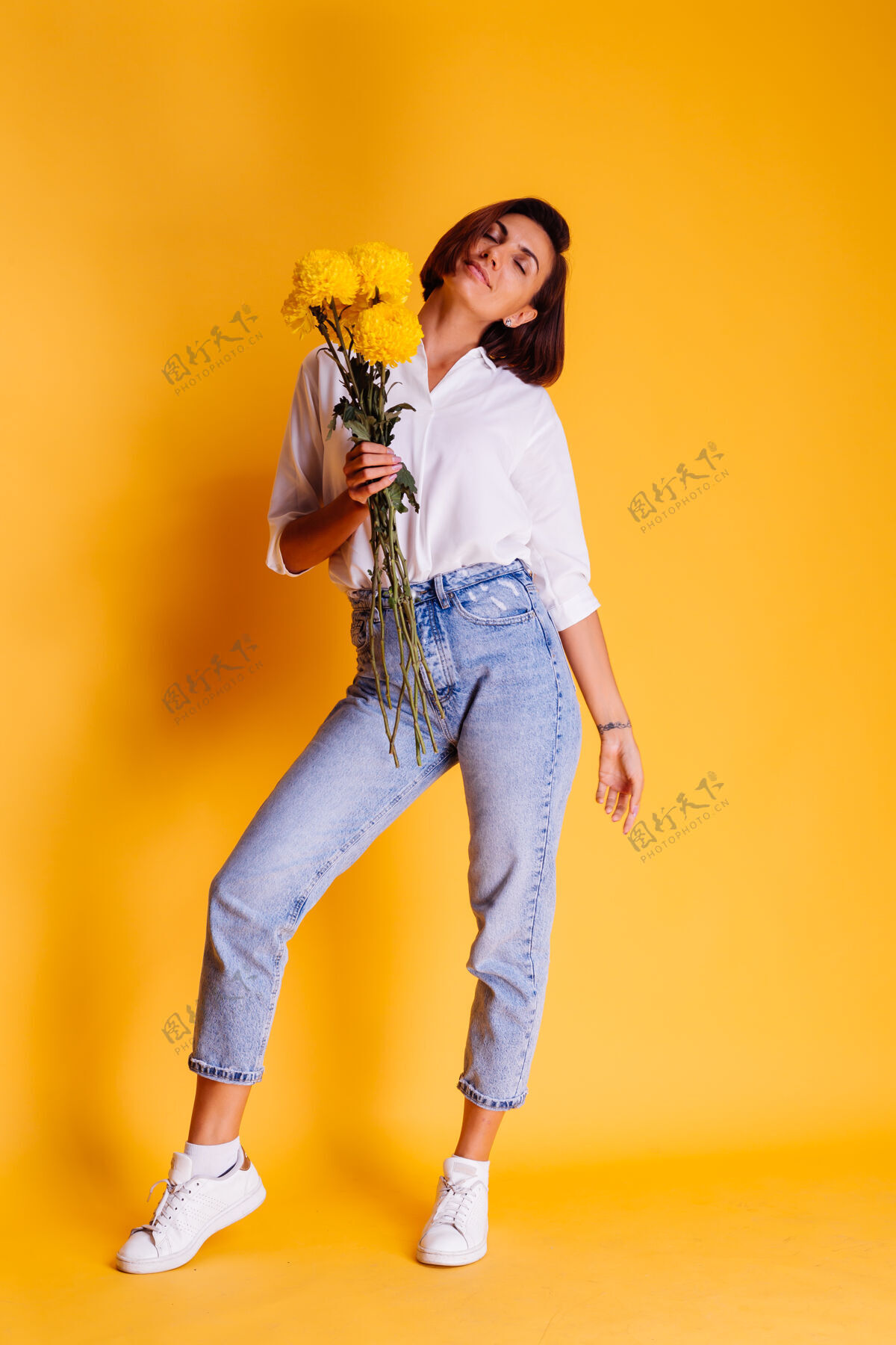 可爱摄影棚拍摄的黄色背景快乐的白人妇女短发穿着休闲服白衬衫和牛仔裤手持一束黄色紫苑惊喜华丽微笑