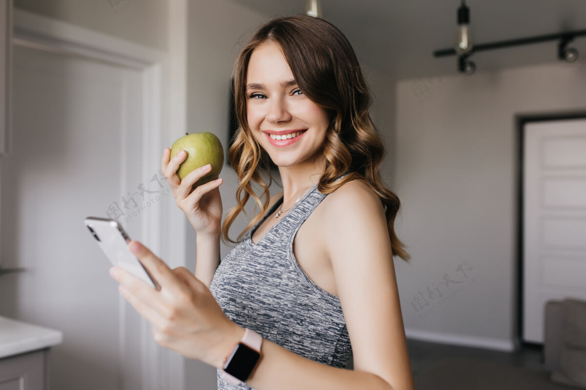 运动漂亮可爱的女孩在家里和青苹果合影幸福的卷发女人微笑着拿着智能手机的室内照片可爱微笑休息