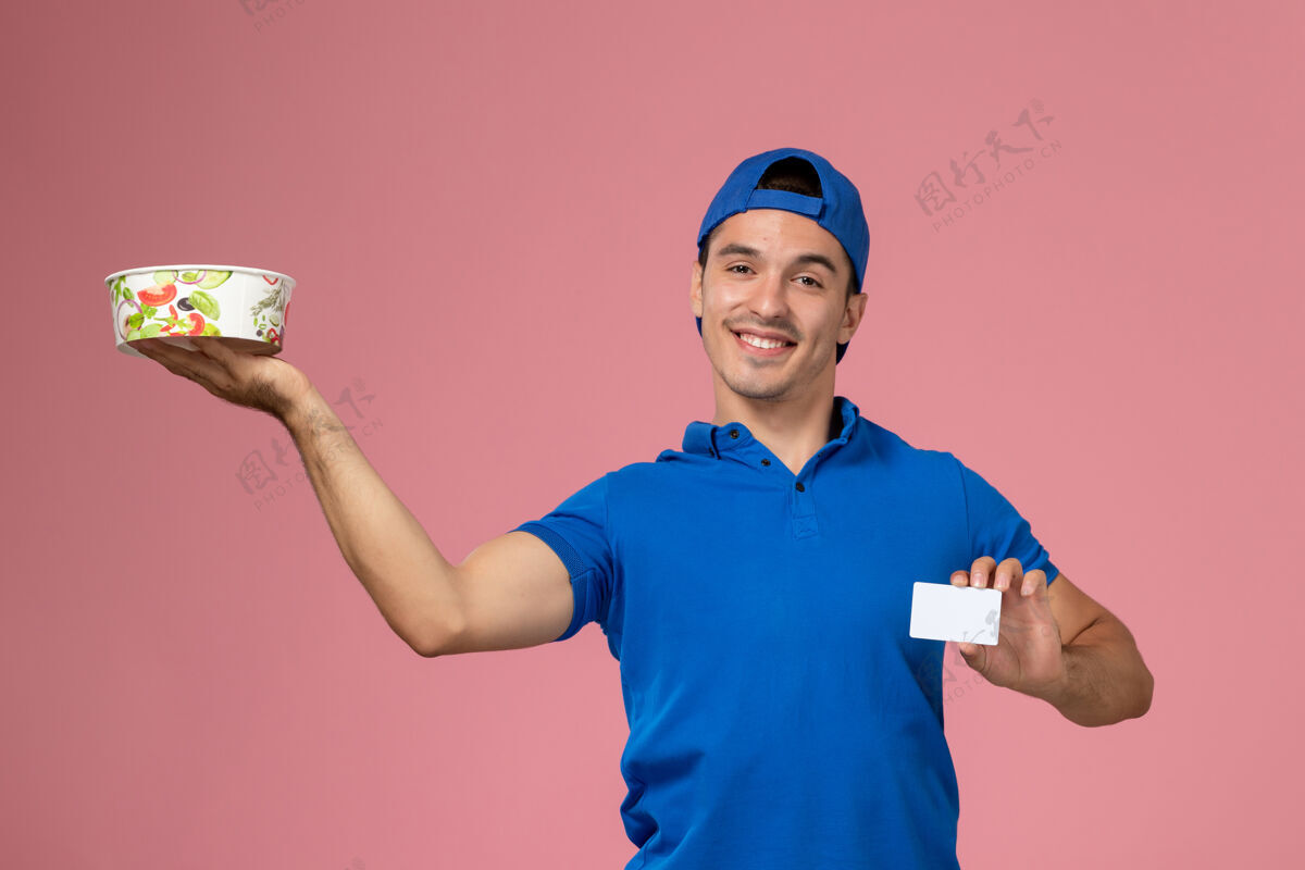 微笑正面图身穿蓝色制服披肩的年轻男性信使手持白色卡片和圆形投递碗 微笑着站在浅粉色的墙上送货碗成人